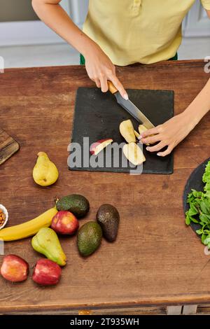 Top-Ansicht der beschnittenen Frau, die Apfel in der Nähe frischer Früchte auf Holztisch schneidet, gesunde pflanzliche Ernährung Stockfoto