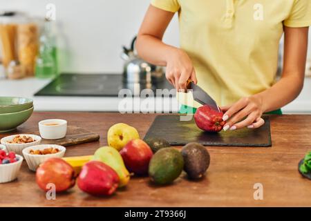 Zugeschnittene Frau schneidet Apfel in der Nähe von frischen Früchten und verschiedenen pflanzlichen Zutaten, pflanzliche Ernährung Stockfoto