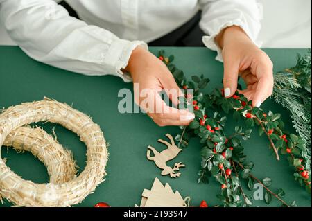 Von unten von der Ernte anonyme Frau, die Weihnachtskranz mit grünen Zweigen mit roten Beeren, grüner Hintergrund neben Kranz Diff Stockfoto