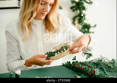 Von unten von der Ernte anonyme Frau, die Weihnachtskranz mit grünen Zweigen mit roten Beeren, grüner Hintergrund neben Kranz Diff Stockfoto