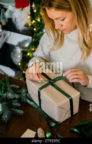 Oben vertikale Ansicht der Frau in Pullover verpackt weihnachtsgeschenk mit Bastelpapier und dekorieren es mit Band auf Tisch legen verschiedene Dekor, Tanne, hinten Stockfoto