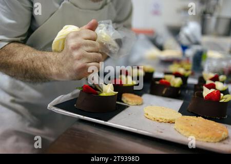 Nahaufnahme der Hände eines leitenden Konditors, der Kuchen mit Früchten in einer Bäckereiküche mit einem Gebäckbeutel dekoriert Stockfoto