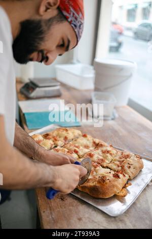 Junger arabischer Pizzamacher in weißem T-Shirt und rotem Hut, der Pizza in Scheiben mit einem Cutter in einer Pizzeria schneidet Stockfoto