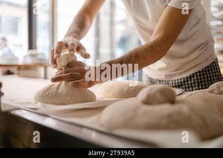 Hände eines nicht erkennbaren männlichen Bäckers in einem weißen T-Shirt, der Brotteig packt und formt, bevor er in den Backofen einer Bäckerei gestellt wird Stockfoto