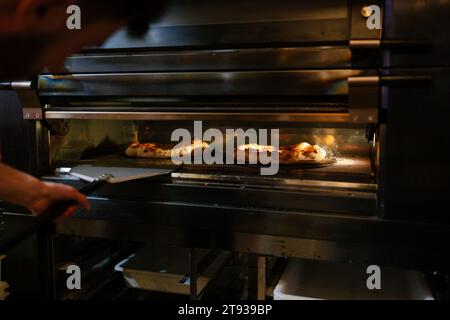 Nicht erkennbarer Pizzamann, der den Zustand von zwei Pizzen im Ofen mit einer Schaufel in einer Pizzeria überprüft Stockfoto