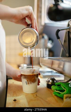 Nahaufnahme der Hände eines nicht erkennbaren Barista, der in einem modernen, spezialisierten Café Kaffee über ein Glas Milch gießt Stockfoto