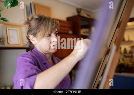 Erwachsene Frau mit blonden Haaren in einem lila Gewand malt mit Pinsel auf Leinwand in ihrem Haus Stockfoto