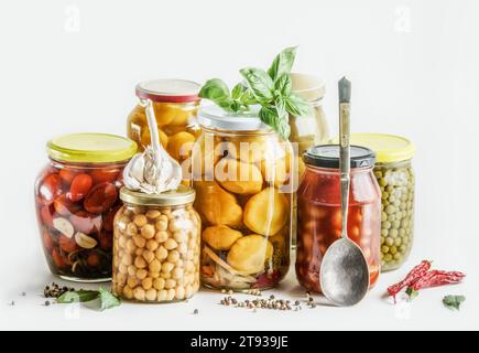 Auswahl an konserviertem Gemüse in Glasgläsern mit Kräutern, Gewürzen und Löffel auf weißem Hintergrund: Konservierte Kichererbsen, Zucchini, Bohnen und Tomaten. Reparieren Stockfoto