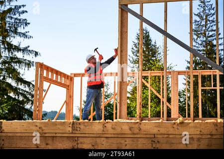Holzrahmenbau wird von einem Zimmermann in der Nähe des Waldes errichtet. Bärtiger Mann mit Brille, der mit Hammer in Nägeln fährt, während er einen Sicherheitshelm und eine Bauweste trägt. Stockfoto