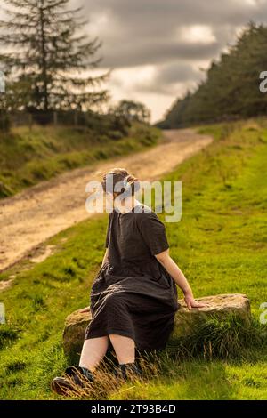 Ein Mädchen im Kleid saß auf einem Sandsteinfelsen und schaute von der Kamera weg auf einem matschigen, nassen Pfad in Osmotherly, North Yorkshire, England, Großbritannien Stockfoto