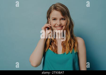 Fröhliche junge Frau mit welligen Haaren lächelnd, berührendes Ohr vor einem weichen blauen Hintergrund Stockfoto