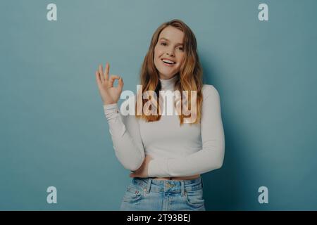 Lächelnde Frau, die mit der Hand OK-Zeichen macht, gekleidet mit weißem Rollkragen und Jeans auf blauem Hintergrund. Stockfoto