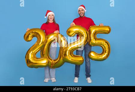 Das Junge, Fröhliche Paar Trägt Santa-Hats Und Hält Goldene 2025 Ballons In Der Hand Stockfoto