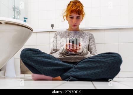 Porträt Teenager Mädchen sitzt auf der Toilette auf dem Boden mit Telefon in der Hand Stockfoto