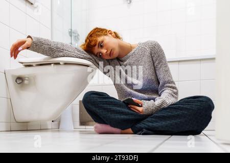 Porträt Teenager Mädchen sitzt auf der Toilette auf dem Boden mit Telefon in der Hand Stockfoto