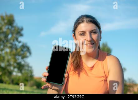 Fröhliche junge Frau, die ihr Handy im Park und schönen blauen Himmel fotografiert Stockfoto