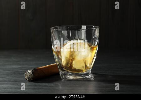 Glas Whiskey mit Eiswürfeln und schwelender Zigarre auf schwarzem Holztisch, Nahaufnahme Stockfoto
