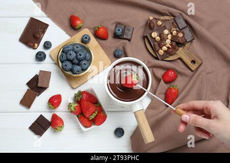 Frau taucht frische Erdbeere in Fonduekanne mit geschmolzener Schokolade an weißem Holztisch, Blick von oben Stockfoto