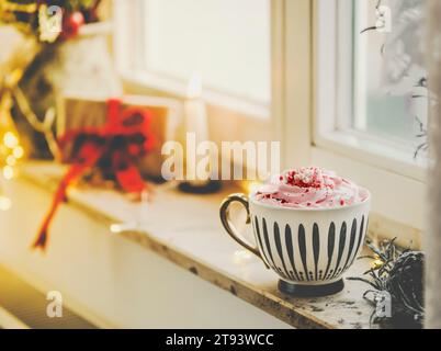 Becher mit heißer Schokolade und Schlagsahne auf Fensterbank mit Kerze, Geschenk und warmem Licht. Hygge überwintert zu Hause mit wärmenden Getränken. Stockfoto