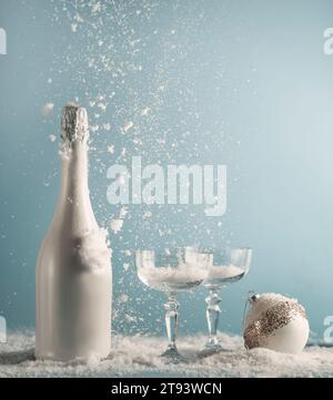 Weiße Champagner-Flasche und Gläser mit Weihnachtskugel auf dem Tisch mit Schneefall auf blauem Wandhintergrund. New Years Konzept mit Getränk und fallendem Schnee Stockfoto