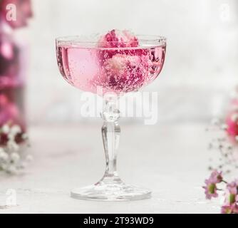 Nahaufnahme eines funkelnden rosafarbenen Getränks mit Rosenblättern in Sektglas am weißen Tisch mit verschwommenem Hintergrund. Aperitif mit eleganter Dekoration für Cele Stockfoto