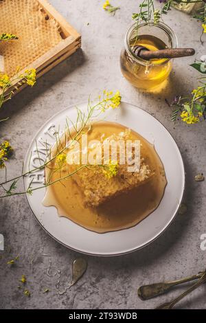 Ganze rohe Honigwabe auf dem Tisch mit Honigglas und hölzerner Dipper, gelbe Blüten mit Blütenblättern. Gesunder und natürlicher flüssiger Süßstoff auf grauem Betonkitch Stockfoto