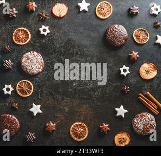 Rahmen mit saisonalen Weihnachtssüßen und aromatischen Gewürzen: Kekse und Lebkuchen, getrocknete Orangenscheiben, Zimt und Sternanis auf dunklem Hintergrund. T Stockfoto