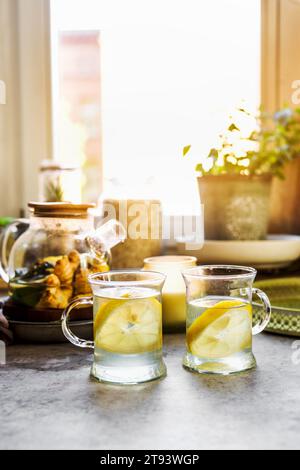 Gesundes Getränk mit Zitrone und Ingwer in Glas Teetassen am Küchentisch mit Tassen, Kerze und Kräutern am Fensterhintergrund mit natürlichem Licht. Erwärmung Stockfoto