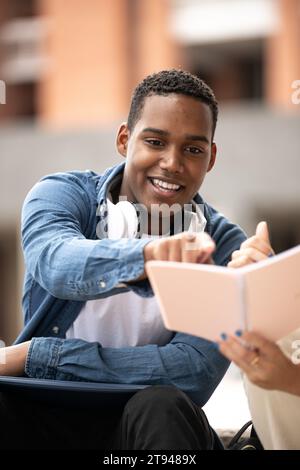 Junger gutaussehender Kerl, der Kopfhörer trägt und auf ein Notebook zeigt. Selbstbewusster Schüler sitzt und liest Notizen mit einem Kollegen. Stockfoto