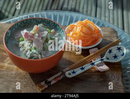 Heißer gekochter Reis oder Reisbrei mit Schweinerippchen Suppe und Schinken serviert mit eingelegten Karotten und Rettich auf hölzernem Schneidebrett. Gesundes Frühstück Stockfoto