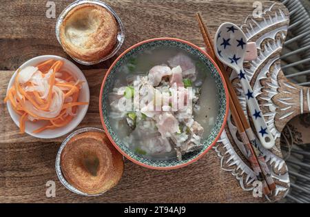 Heißer gekochter Reis oder Reisbrei mit Schweinerippchen Suppe und Schinken serviert mit frittiertem Teig, eingelegter Karotte und Rettich auf hölzernem Schneidebrett. Er Stockfoto