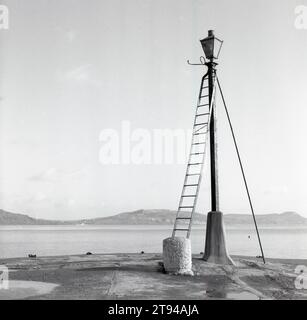 1950er Jahre, historisch, eine gusseiserne traditionelle Gas-beleuchtete Laterne mit Betonsockel am Rand eines Hafens mit Blick auf das Wasser, mit angebrachten Metallleitern, verwendet, um sie hinaufzuklettern und zu belüften. Hier sind die Leitern dauerhaft am Lampenpfosten befestigt, während in einer Stadt der lamplighter seine eigenen trug. Die Gasbeleuchtung wurde 1792 von William Murdoch erfunden und war sehr pflegeleicht, da die Lichter jede Nacht manuell angezündet und jeden Morgen gelöscht werden mussten und mit der Erfindung der Elektrizität durch elektrische Glühbirnen ersetzt wurden. Stockfoto