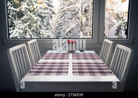 Winterfenster und Küchentisch zum Essen. Inneneinrichtung und Möbel. Schnee in Bäumen im Wald im Hintergrund. Skandinavisches Haus. Stockfoto