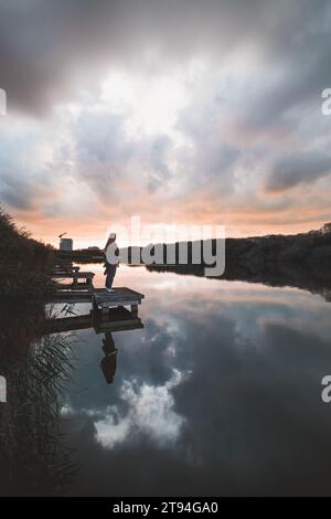 Dame im Alter von 20-25 Jahren, die auf einem hölzernen Pier an einem Teich stand und den Sonnenuntergang über der Stadt Oostende im Westen Belgiens beobachtete. Dramatischer rot-oranger Himmel. Stockfoto