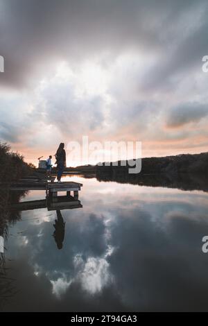 Dame im Alter von 20-25 Jahren, die auf einem hölzernen Pier an einem Teich stand und den Sonnenuntergang über der Stadt Oostende im Westen Belgiens beobachtete. Dramatischer rot-oranger Himmel. Stockfoto