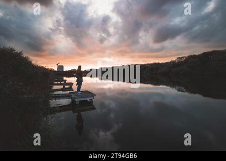 Dame im Alter von 20-25 Jahren, die auf einem hölzernen Pier an einem Teich stand und den Sonnenuntergang über der Stadt Oostende im Westen Belgiens beobachtete. Dramatischer rot-oranger Himmel. Stockfoto