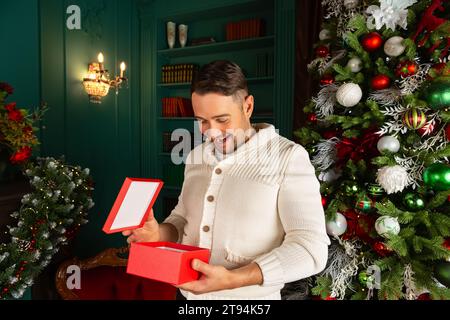 Aufgeregter junger Mann, der ein Weihnachtsgeschenk zu Hause eröffnet. Porträt des glücklichen, schönen emotionalen Mannes, der die Weihnachtsgeschenkbox öffnet. Stockfoto