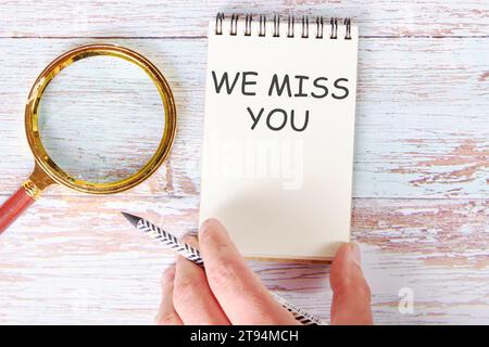 WIR VERMISSEN SIE auf einem leeren Blatt eines Notizbuchs, das von einer Hand eines Mannes mit einem Bleistift berührt wird, und daneben liegt eine Lupe Stockfoto