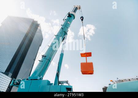 Ein großer blauer LKW-Kran steht auf einem Gelände in der Nähe eines großen modernen Gebäudes betriebsbereit. Der größte Lkw-Kran mit gelber Wiege zum Lösen von c Stockfoto