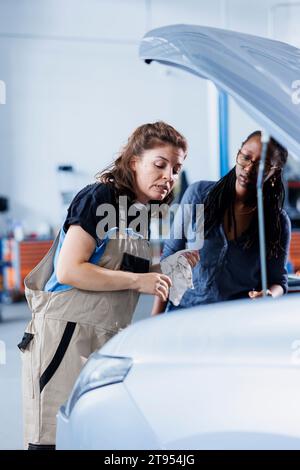 Ingenieur in der Garage, der das Fahrzeug für BIPOC Frau repariert und unter der Motorhaube schaut, um verbleibende Ölleckagen zu entfernen. Der Mitarbeiter führt routinemäßige Reinigung der Auspuffanlage am Kundenfahrzeug durch Stockfoto