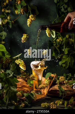 Frau gießt Bier aus einer Flasche in ein Glas. Komposition mit fliegenden Hopfen und Spritzern Bier. Stockfoto