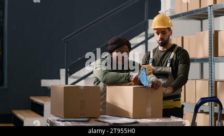 Mann mit Helm im Lager, der Pappkartons an afroamerikanische Manager aushändigt und Pakete versiegelt. Mitarbeiter des Lagerbetriebs sichern Pakete, um Probleme bei der Zustellung zu vermeiden Stockfoto