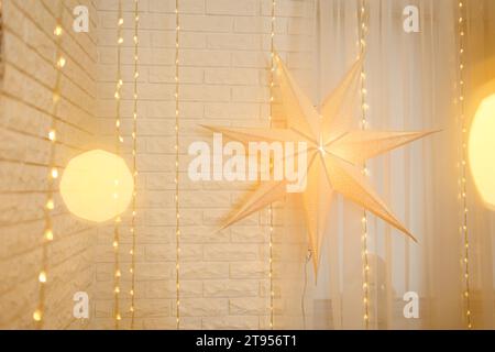 Weihnachtsstern auf goldenem Lichterhintergrund an der Wand im Wohnzimmer. Großer Papierstern leuchtet vor dem Hintergrund der Neujahrsgirlande. Stimmungsvolles fest Stockfoto