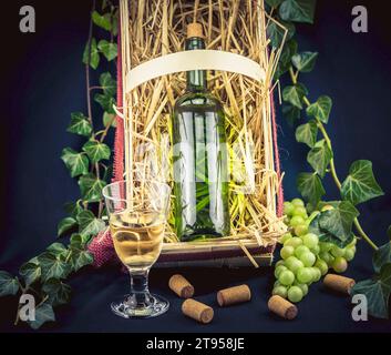 Weinrebe, Rebe (Vitis vinifera), Flasche Weißwein in einem Strohkasten, Glas Weißwein umgeben von Efeu und grünen Trauben Stockfoto