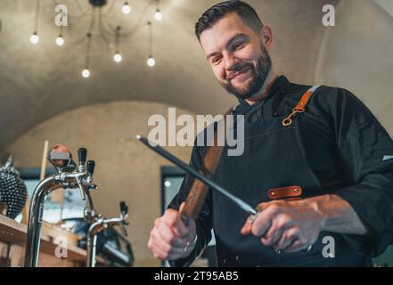 Lächelnder bärtiger Koch in schwarzer Uniform mit Schürzenschärfmesser mit Honing Steel Werkzeug. Berufsberuf, harte Arbeit, Lebensmittelzubereitungen Stockfoto