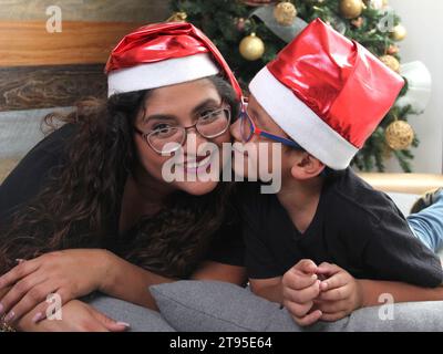 Doppelt doppelt so viele alleinerziehende Mutter und ein 7-jähriger dunkelhäutiger Latino-Sohn tragen Weihnachtsmannshüte vor dem Weihnachtsbaum, um gemeinsam in Einsamkeit zu feiern Stockfoto