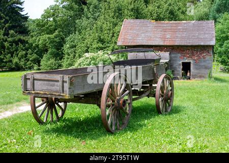 Ein alter Holzwagen auf dem Gras in einer ländlichen Umgebung mit einer Scheune im Hintergrund Stockfoto