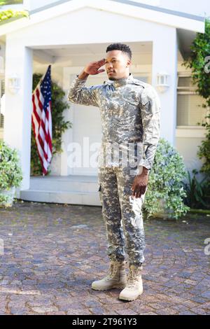 Fokussierter afroamerikanischer männlicher Soldat, der vor dem Haus steht und mit us-Flagge grüßt, Kopierraum Stockfoto