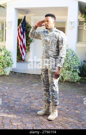 Fokussierter afroamerikanischer männlicher Soldat, der vor dem Haus steht und mit us-Flagge grüßt, Kopierraum Stockfoto