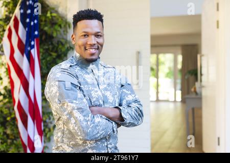 Männlicher afroamerikanischer Soldat, der mit Armen vor dem Haus steht, mit us-Flagge, Kopierraum Stockfoto